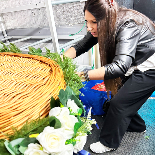 Donna, owner of Orchid Memories, standing beside a wicker coffin, carefully dressing it with a garland of silk flowers.
